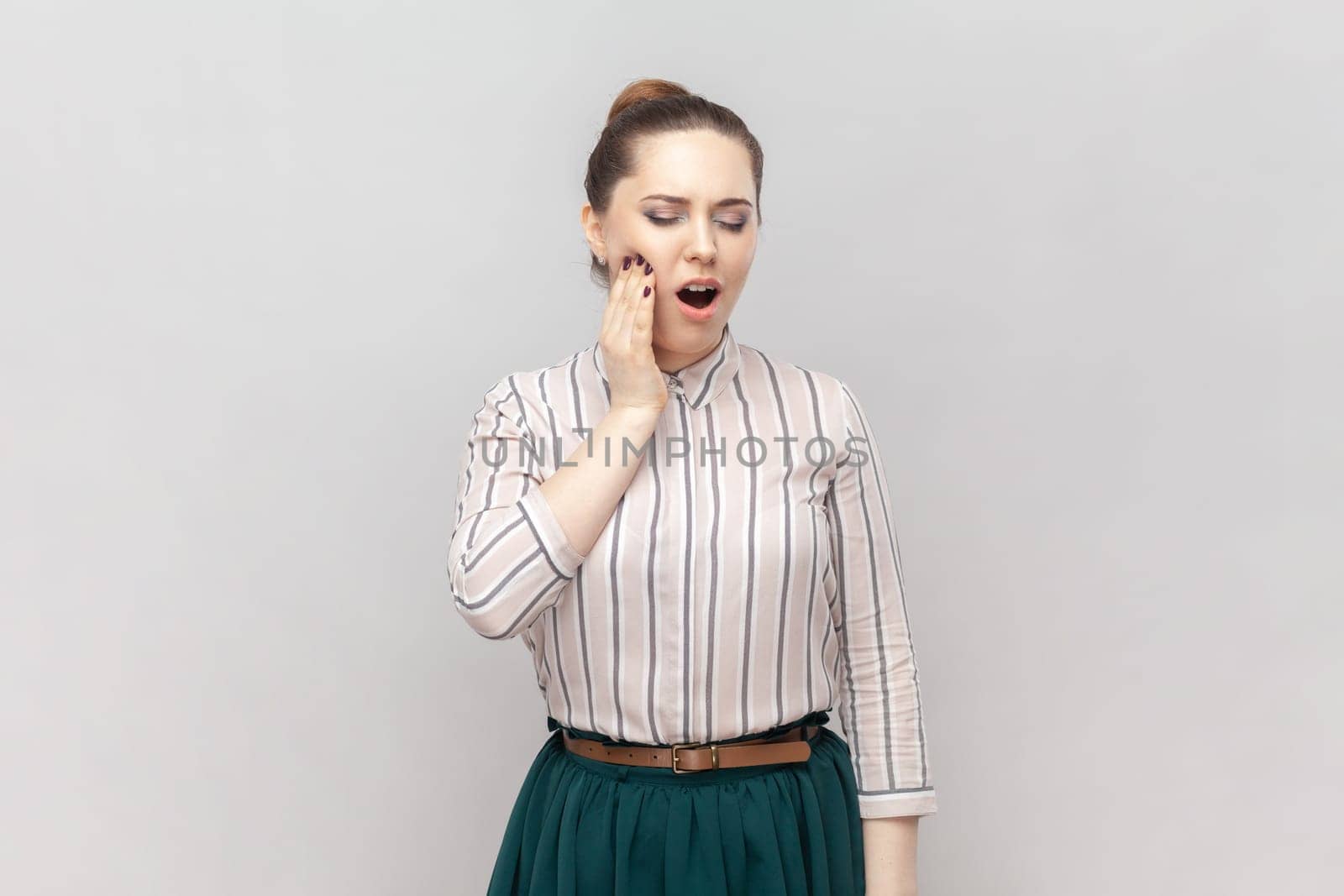 Portrait of sick unhealthy ill woman wearing striped shirt and green skirt standing touching her painful cheek, suffering terrible toothache. Indoor studio shot isolated on gray background.