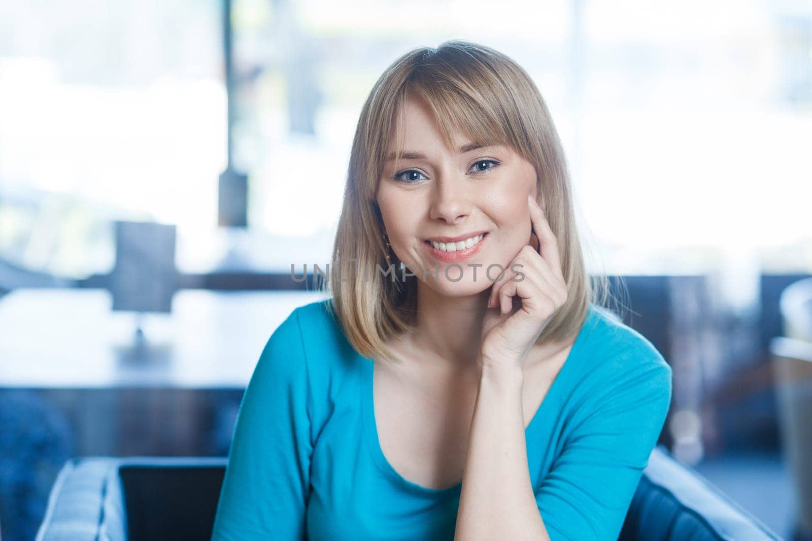 Portrait of smiling happy satisfied young woman with blonde hair in blue shirt sitting alone, looking at camera with toothy smile, being in good mood. Indoor shot in cafe with big window on background