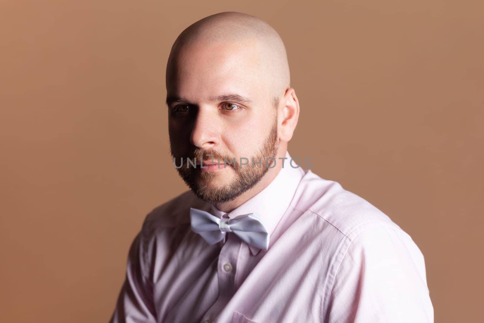 Portrait of handsome attractive bald bearded man looking away with pensive thoughtful facial expression, wearing light pink shirt and bow tie. Indoor studio shot isolated on brown background.