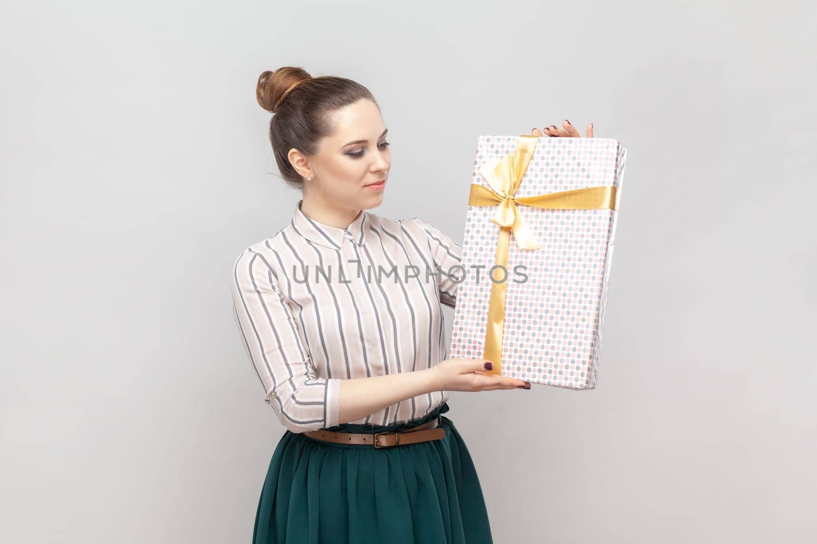 Portrait of young adult beautiful woman wearing striped shirt and green skirt standing showing wrapped present box, congratulating with holiday. Indoor studio shot isolated on gray background.