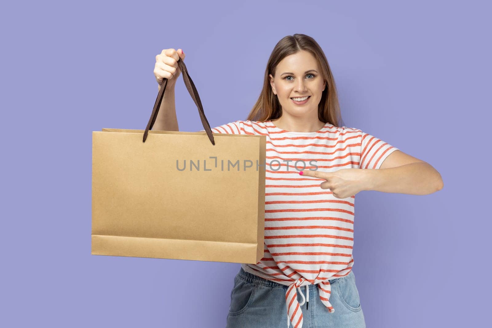 Portrait of attractive positive young blond woman wearing striped T-shirt standing pointing at shopping bag, looking at camera with toothy smile. Indoor studio shot isolated on purple background.
