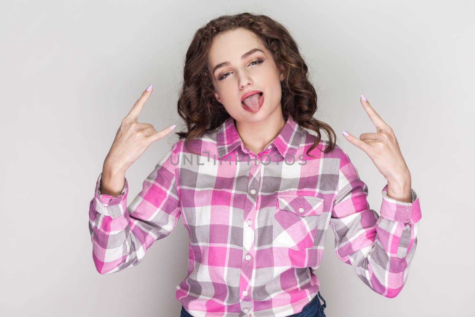 Portrait of energetic young woman with curly hair makes rock n roll gesture, enjoys cool music, showing tongue out, wearing pink checkered shirt. Indoor studio shot isolated on gray background.