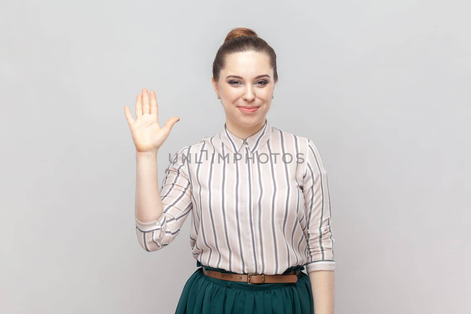 Portrait of pleased pretty positive woman wearing striped shirt and green skirt standing waving hand, saying hello or bye, having meeting. Indoor studio shot isolated on gray background.