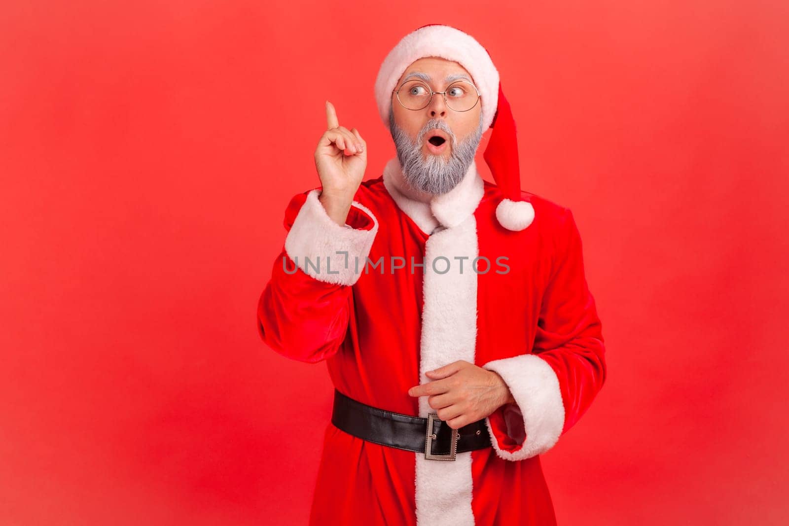 Elderly man with gray beard wearing santa claus costume raising finger and having genius idea of Christmas celebration, looking amazed, creative thought. Indoor studio shot isolated on red background.