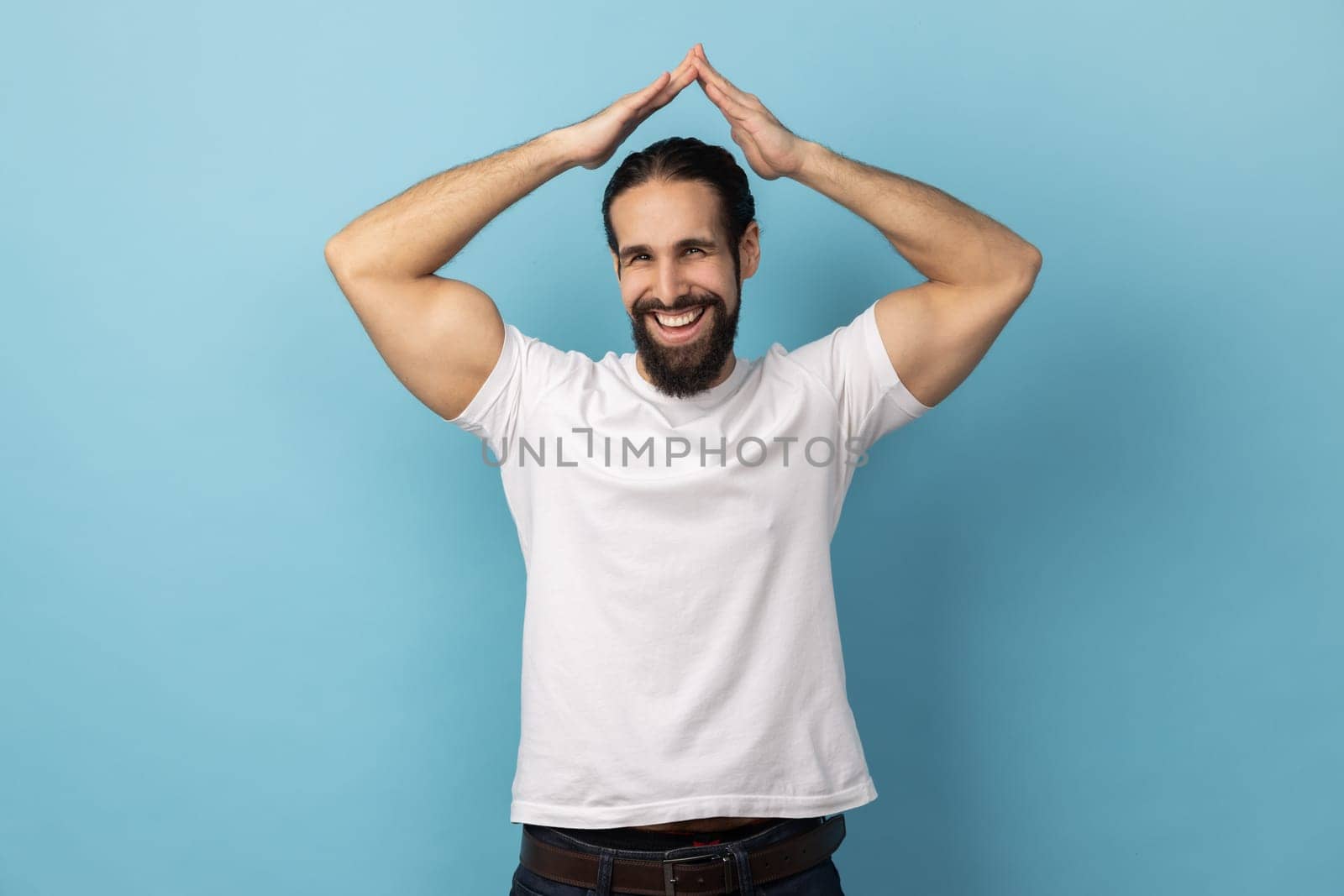 Safety and house insurance. Portrait of man with beard wearing white T-shirt making roof gesture over head and smiling to camera with amiable expression. Indoor studio shot isolated on blue background