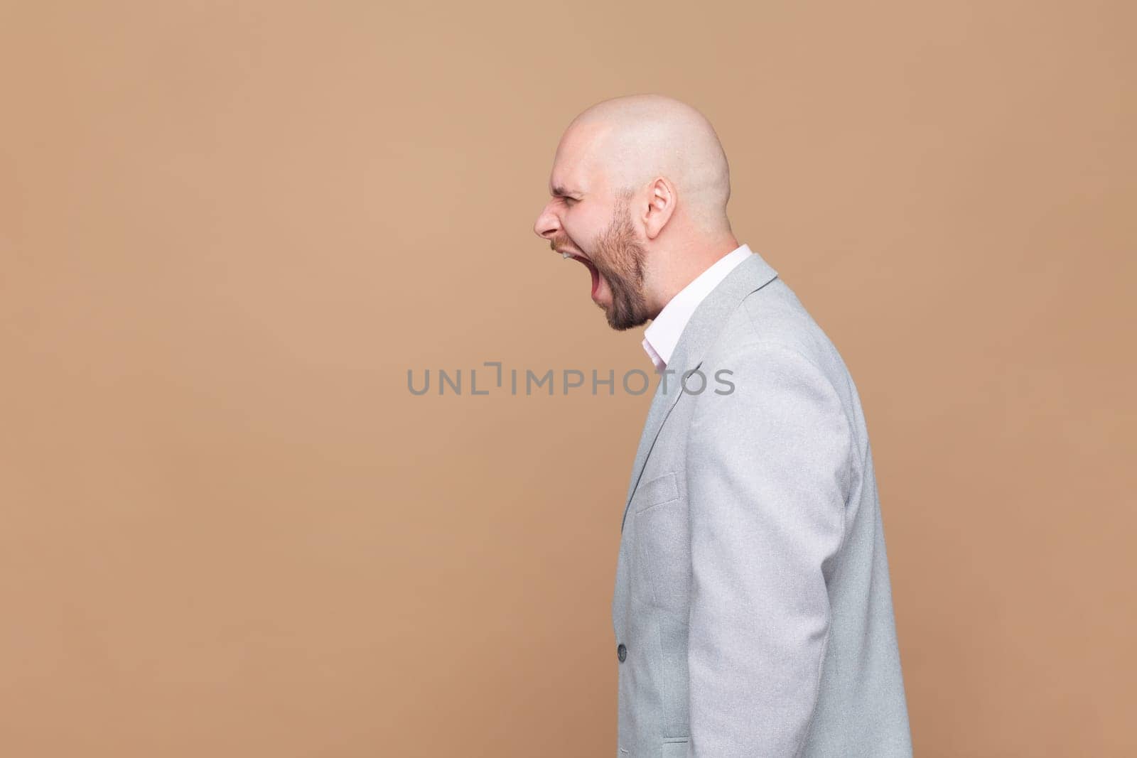 Side view portrait of angry or shocked bald bearded man standing, looking forward and screaming, copy space foe advertisement, wearing gray jacket. Indoor studio shot isolated on brown background.