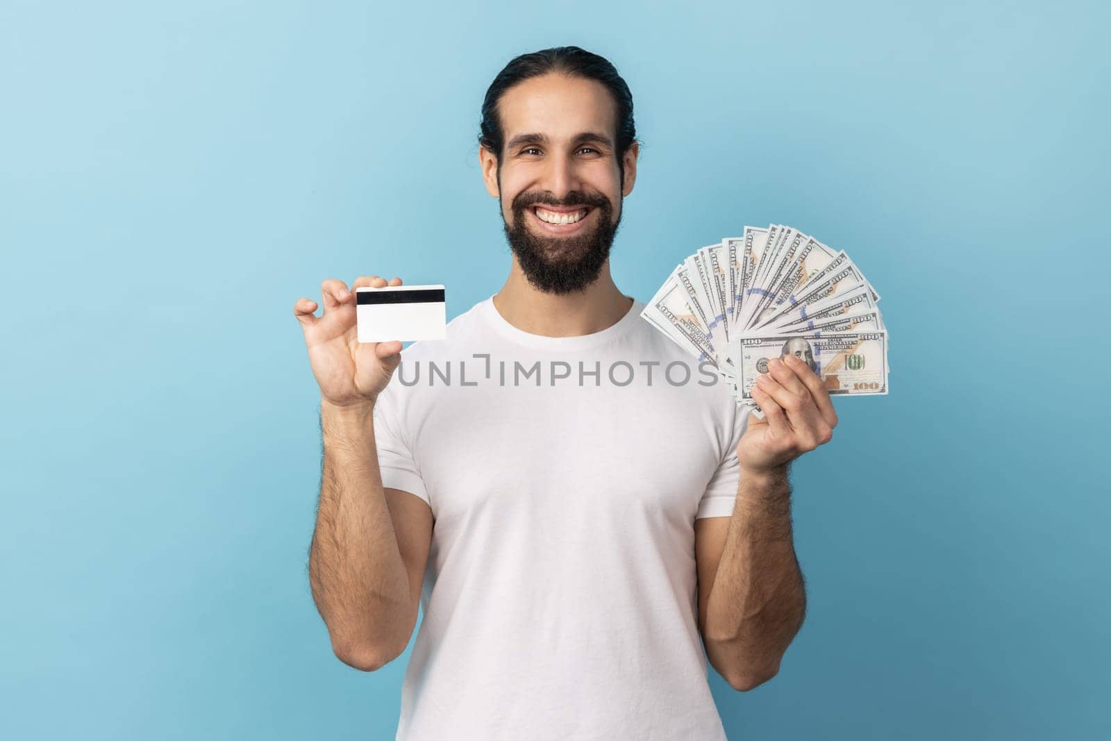 Man with beard wearing white T-shirt holding credit card and dollar banknotes, feeling rich with lot of money, happy to get bank loan for shopping. Indoor studio shot isolated on blue background.