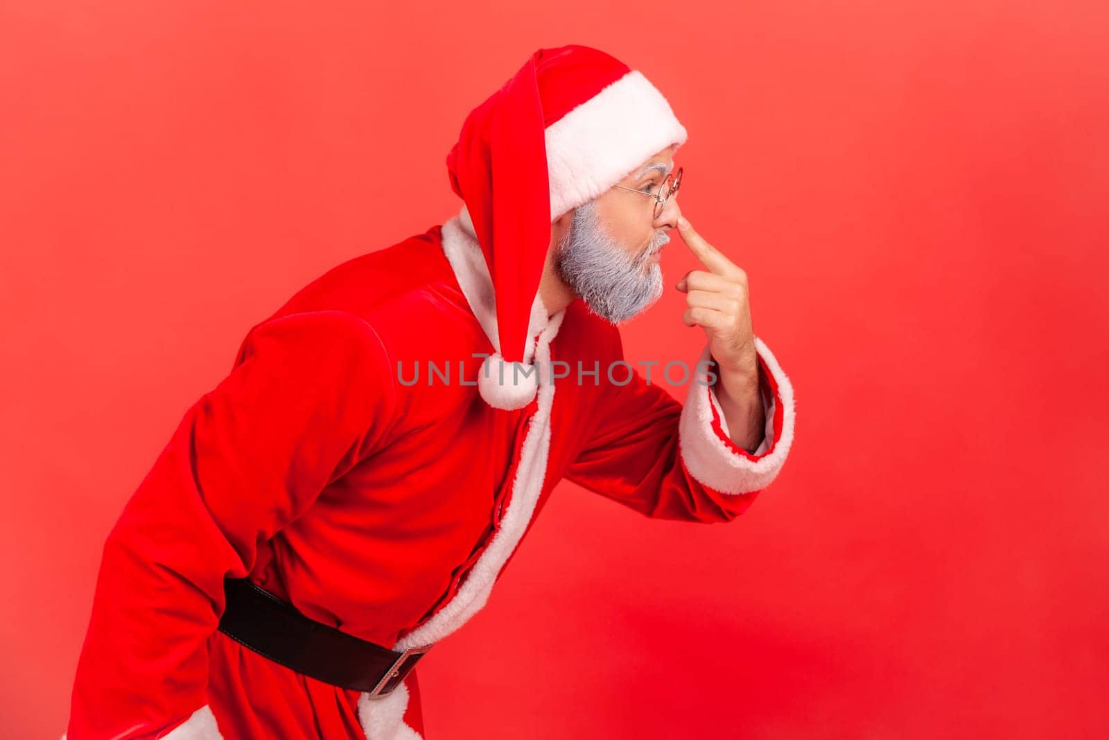 Side view portrait of elderly man with gray beard wearing santa claus costume touching nose with finger, reproaching liar in deception. Indoor studio shot isolated on red background.