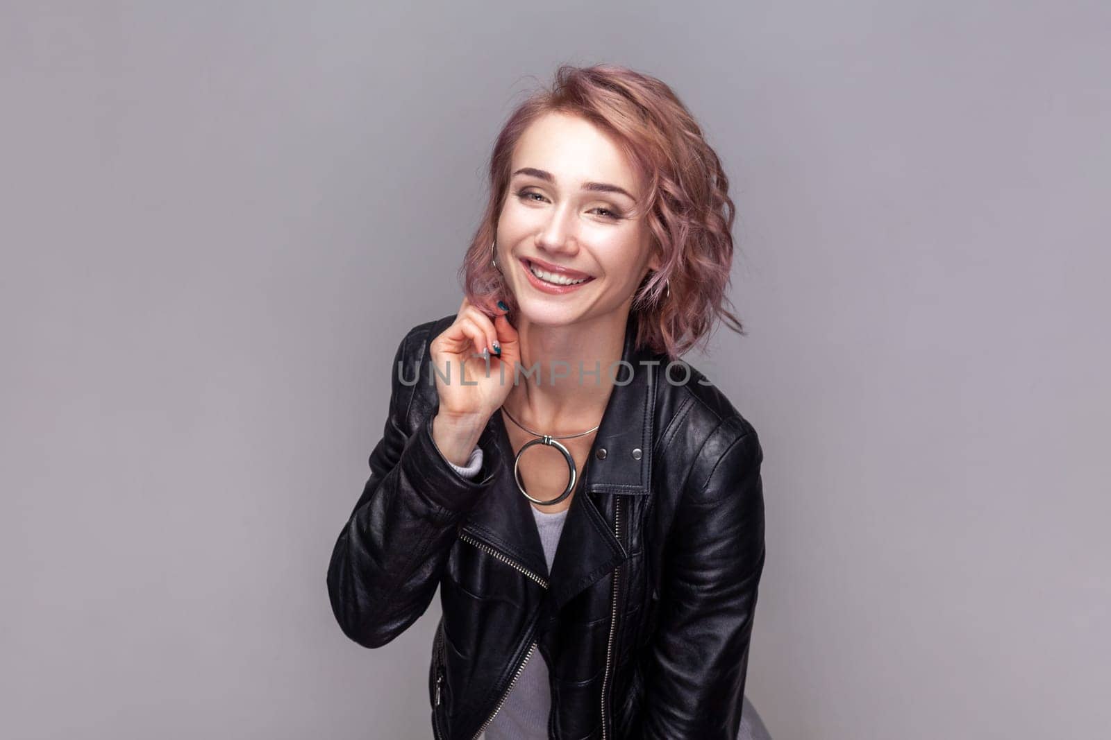 Portrait of charming beautiful adorable woman with short hairstyle standing looking at camera with toothy smile, wearing black leather jacket. Indoor studio shot isolated on grey background.