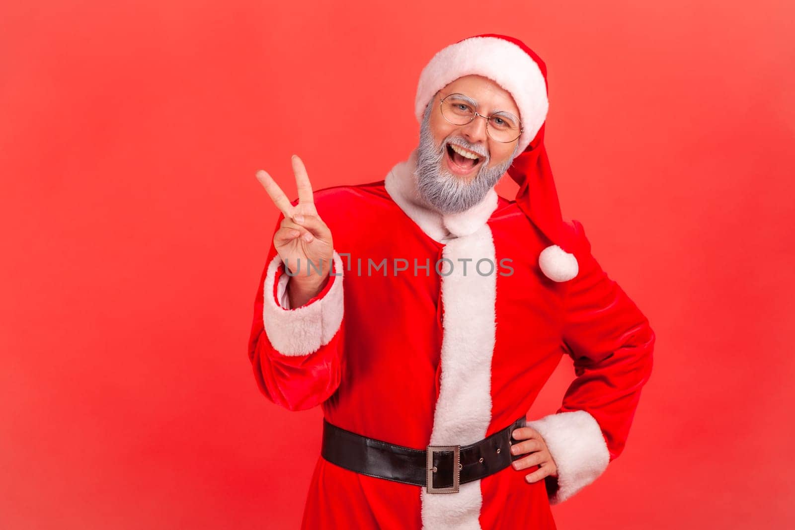 Amazed elderly man with gray beard wearing santa claus costume standing with raised hands and showing v sign or peace, victory gesture. by Khosro1