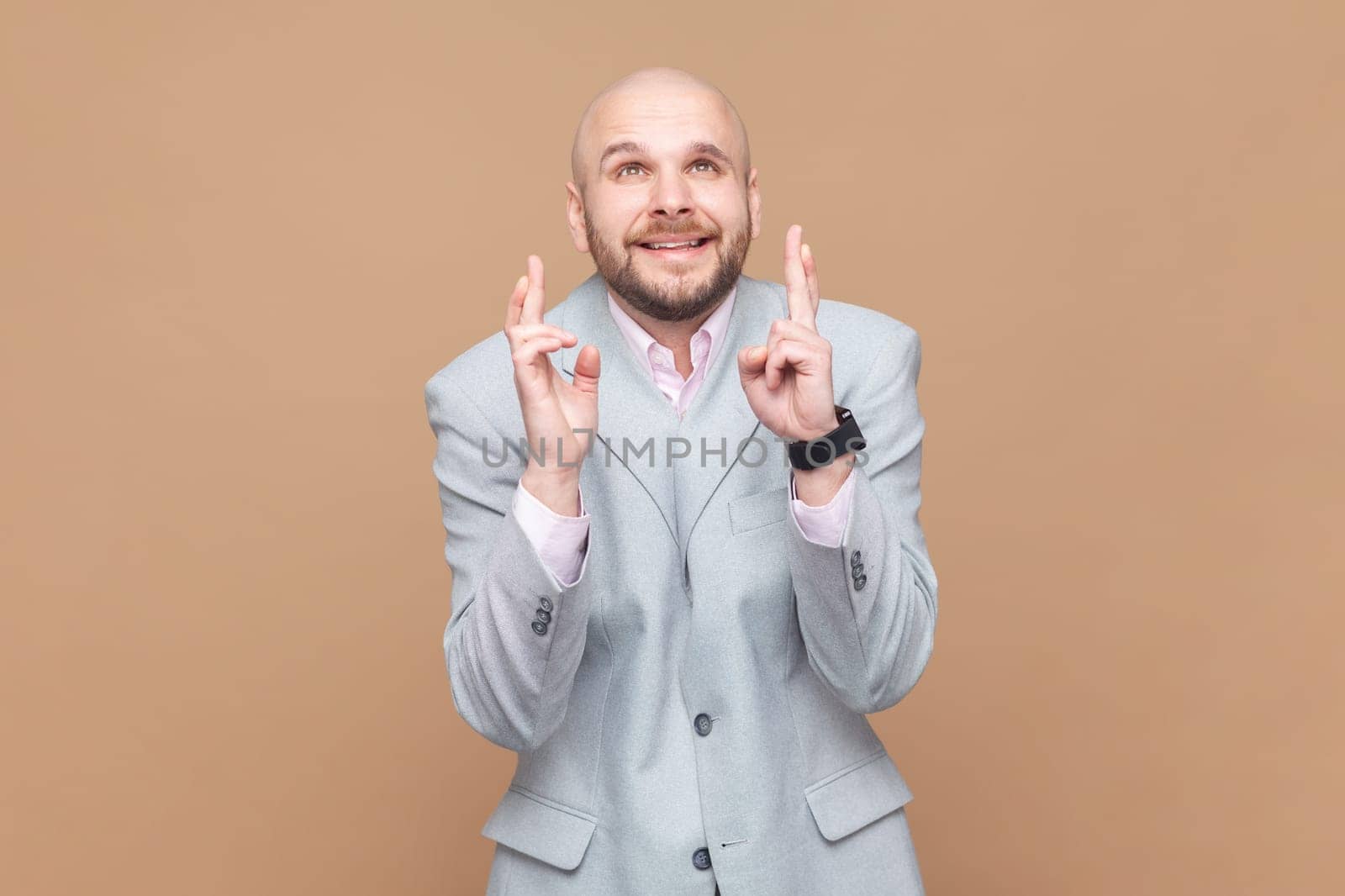 Portrait of hopeful nervous bald bearded man crossing fingers for luck, hope for better, ritual, looks up with hopeful look, wearing gray jacket. Indoor studio shot isolated on brown background.