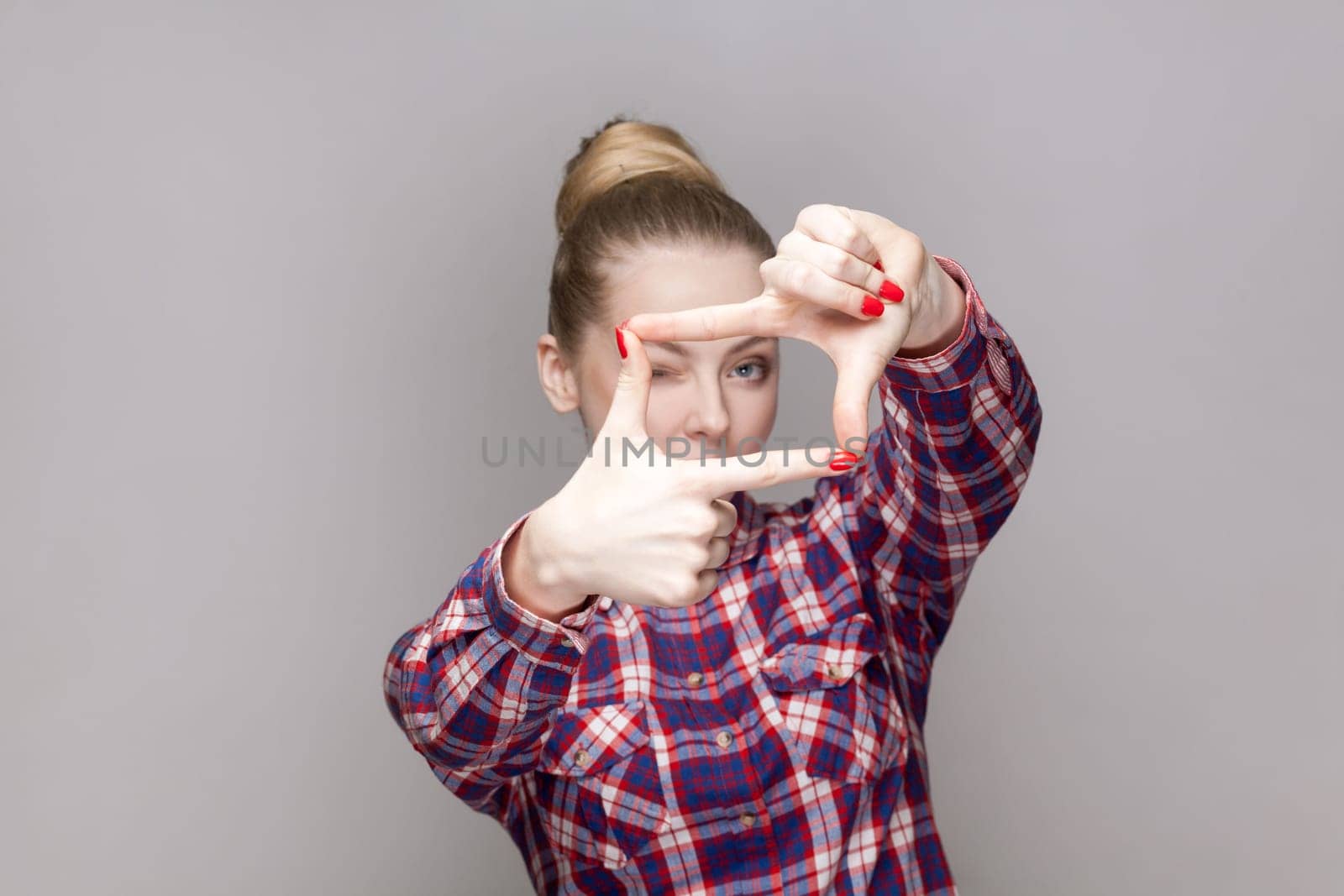Portrait of attentive concentrated woman photographer with bun hairstyle looking at camera through frame of fingers, wearing checkered shirt. Indoor studio shot isolated on gray background.