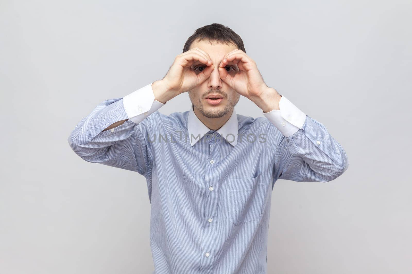 Portrait of curious funny young adult man standing hands near eyes, making binocular gesture, looking at something interesting, wearing light blue shirt. Indoor studio shot isolated on gray background