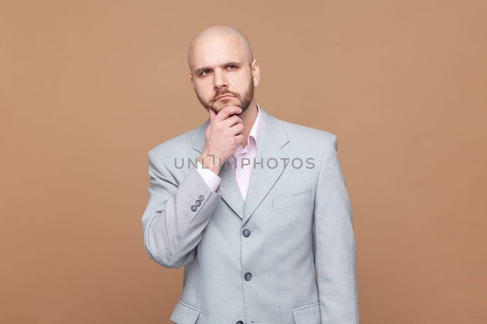 Let me think about it. Portrait of thoughtful handsome bald bearded man holds chin and looks pensively away tries to solve problem, wearing gray jacket. Indoor studio shot isolated on brown background