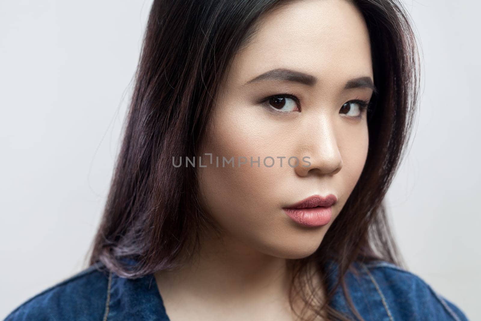 Closeup portrait of serious concentrated beautiful young adult brunette woman with makeup and long hair, looking at camera. Indoor studio shot isolated on gray background.