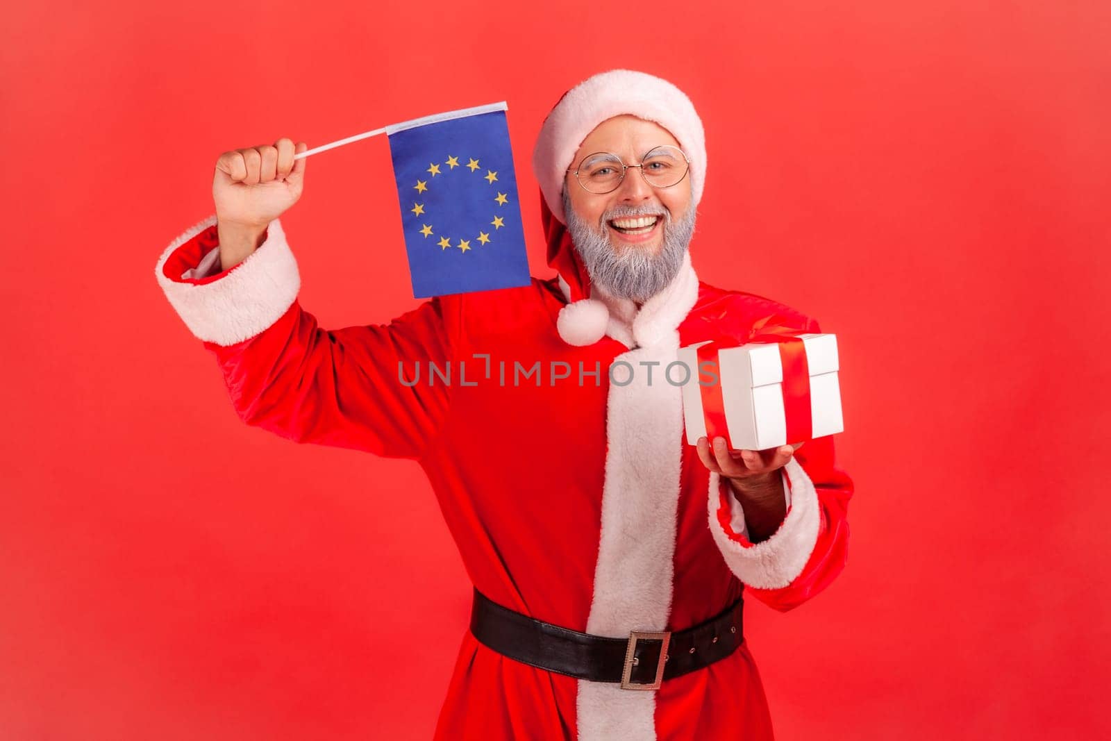 Portrait of smiling elderly man with gray beard wearing santa claus costume holding wrapped present box and waving Europe union flag, looks at camera. by Khosro1