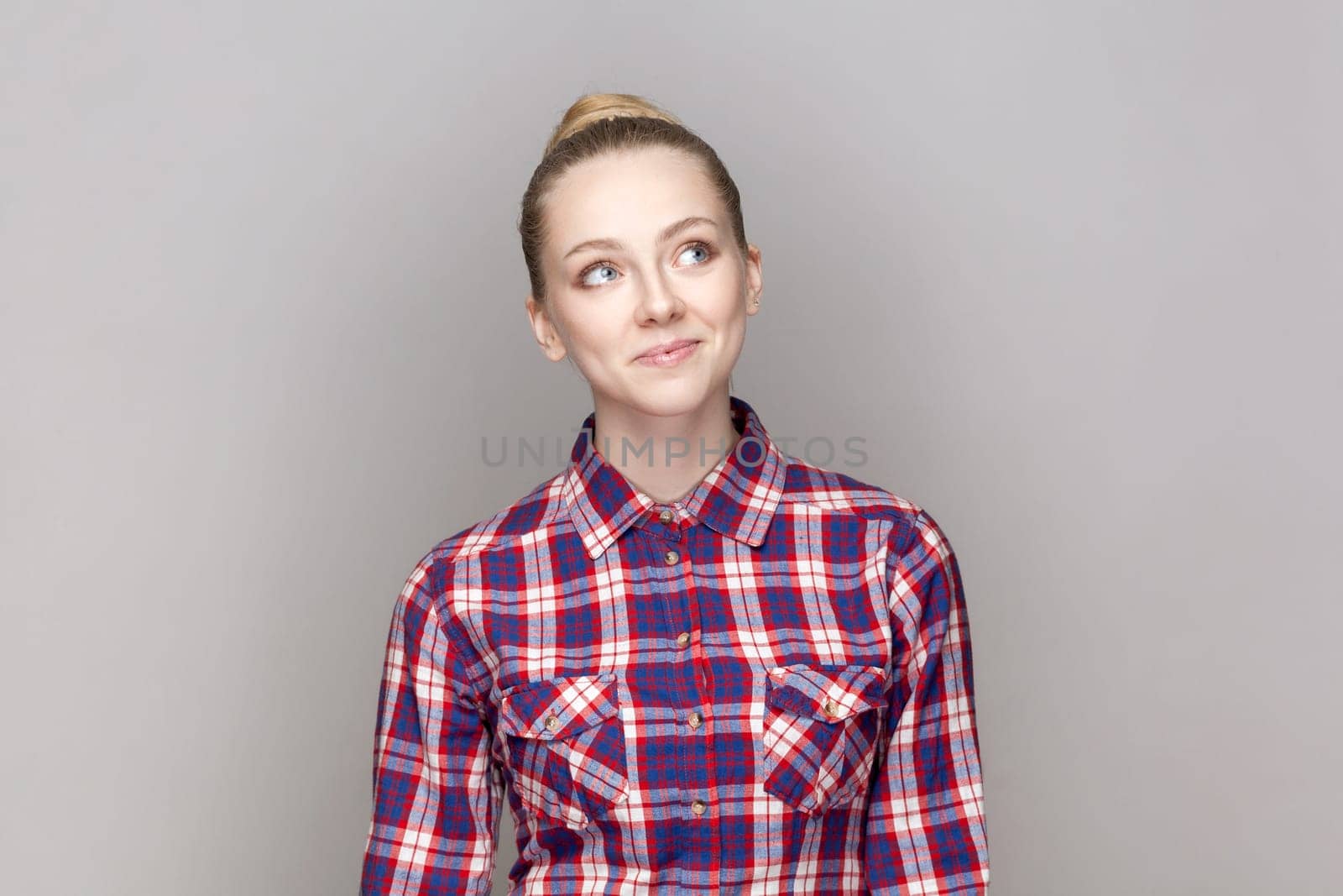 Portrait of positive smiling adorable woman with bun hairstyle looking away, thinking about future, dreaming, wearing checkered shirt. Indoor studio shot isolated on gray background.
