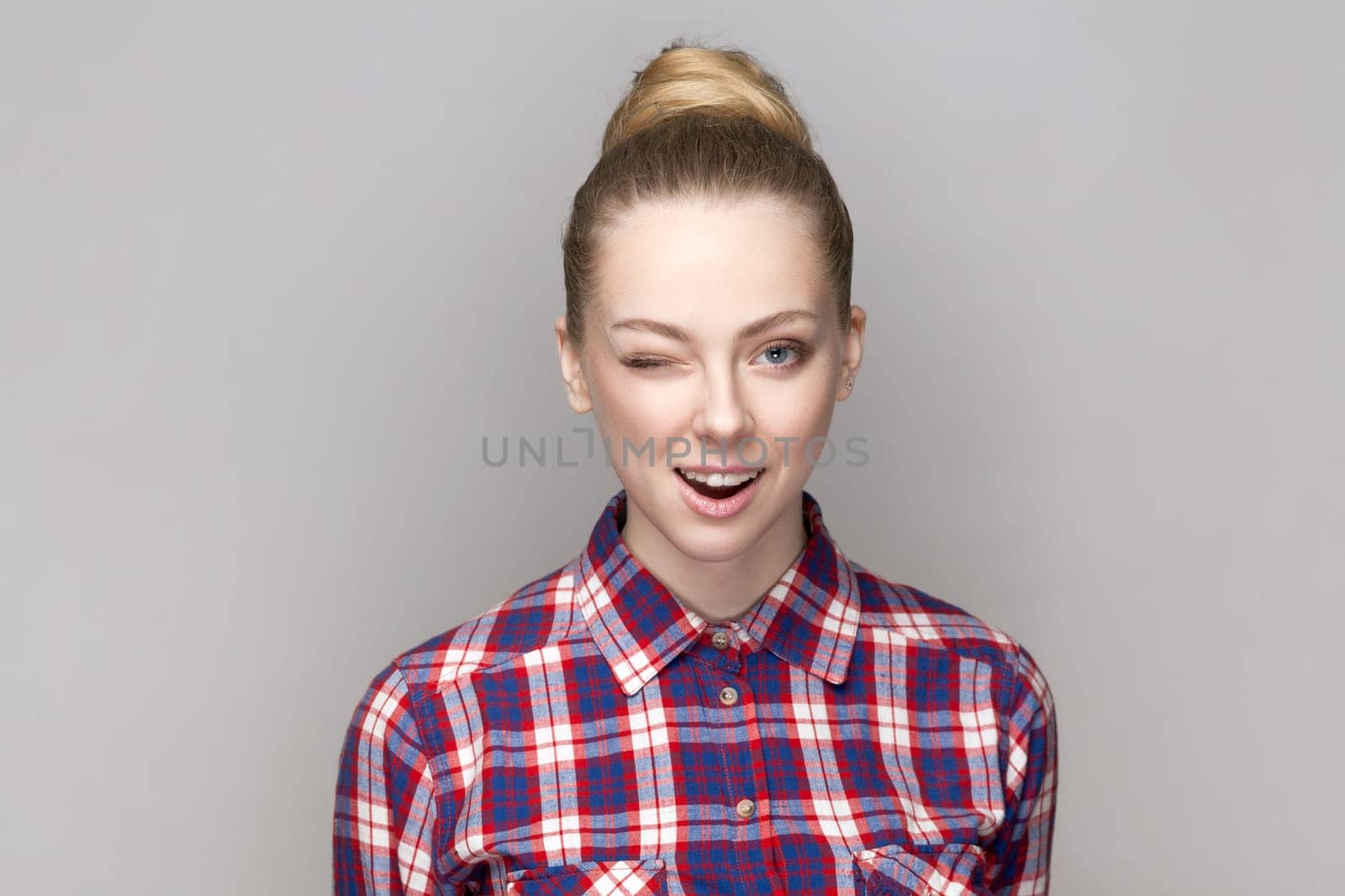 Portrait of flirting positive adorable woman with bun hairstyle standing blinking her eye, looking at camera, winking, wearing checkered shirt. Indoor studio shot isolated on gray background.