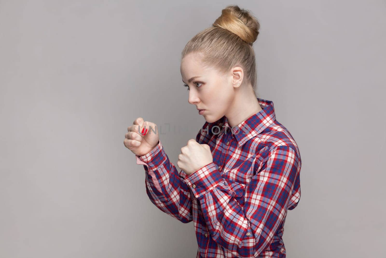 Side view portrait of angry woman with bun hairstyle standing with clenched fists, being ready to attack somebody, wearing checkered shirt. Indoor studio shot isolated on gray background.