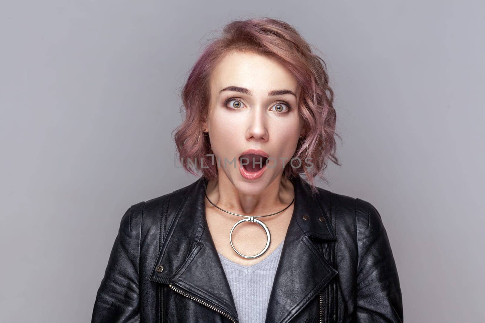 Portrait of shocked amazed surprised woman with short hairstyle standing looking at camera with big eyes and open mouth, wearing black leather jacket. Indoor studio shot isolated on grey background.