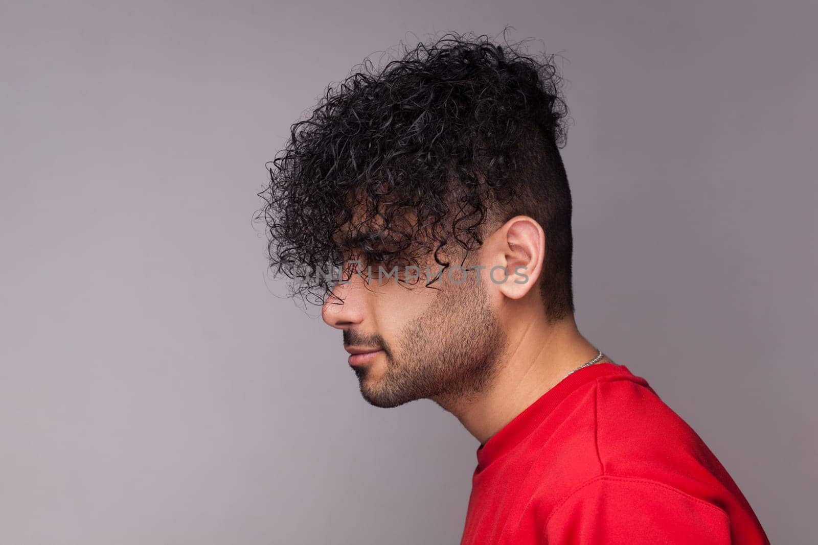 Side view portrait of attractive handsome young adult bearded man with curly hair, wearing red jumper, looking ahead, showing his hairdo. Indoor studio shot isolated on gray background.