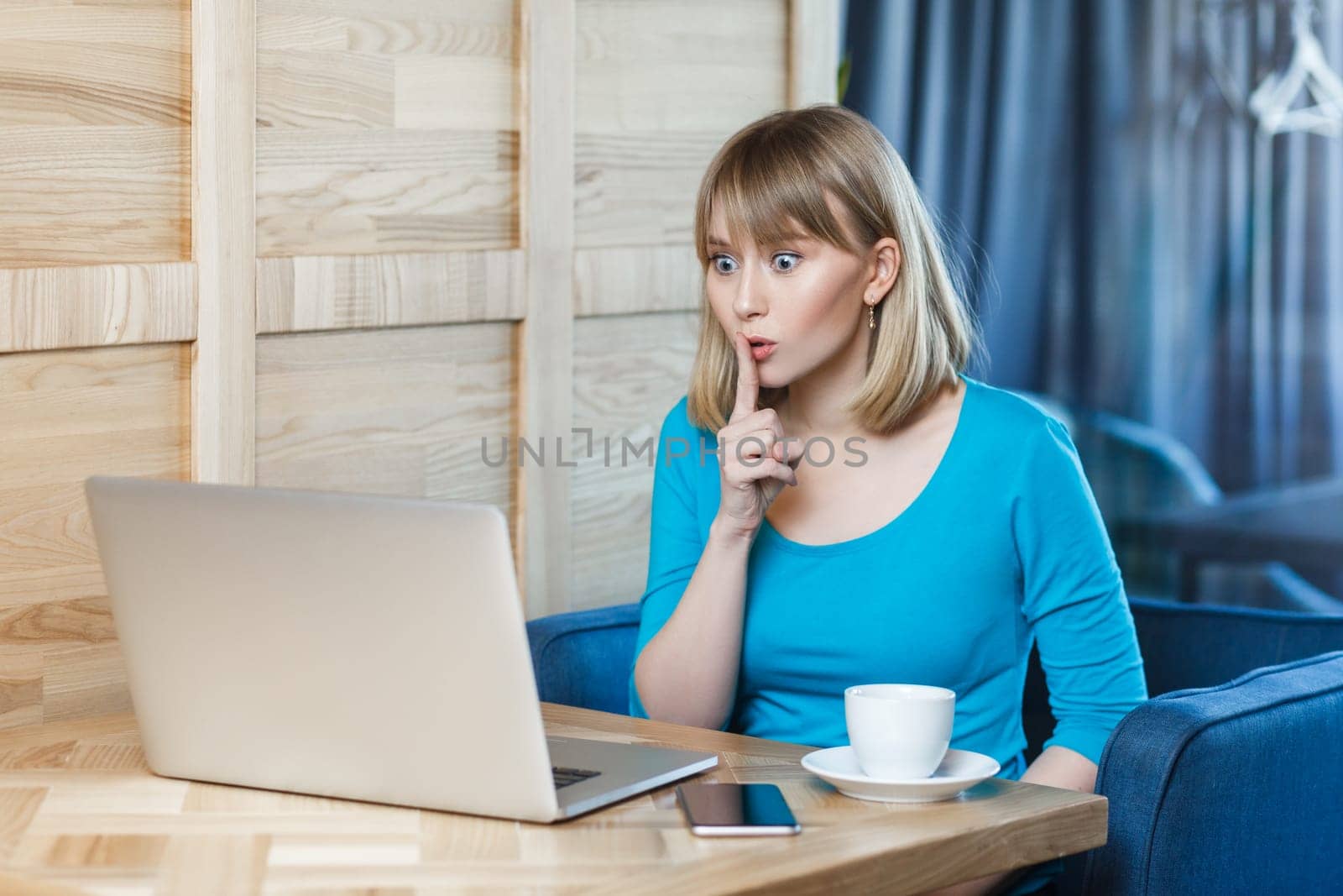 Portrait of serious adorable young woman with blonde hair in blue shirt sitting at table and working on laptop, has video call, showing shh gesture. Indoor shot, cafe background.
