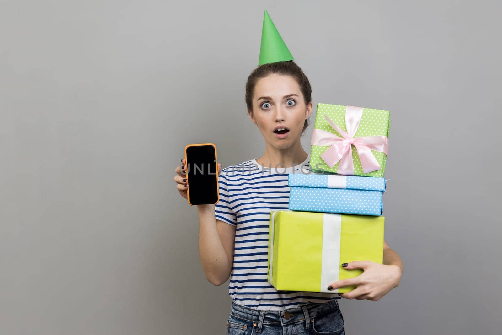 Portrait of shocked woman wearing striped T-shirt and party cone, holding stack of present boxes and showing mobile phone with blank display. Indoor studio shot isolated on gray background.