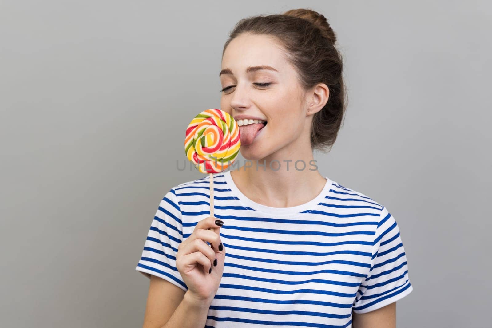 Portrait of happy woman wearing striped T-shirt standing with tongue out, dreaming and tempted to taste lollipop, round sweet sugary rainbow candy. Indoor studio shot isolated on gray background.