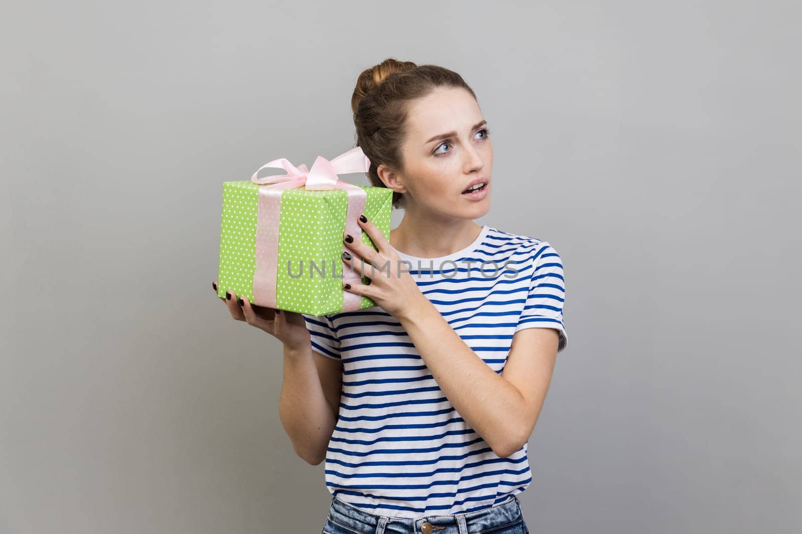 Portrait of woman wearing striped T-shirt shaking wrapped present box, being interested what inside, looking away with curious facial expression. Indoor studio shot isolated on gray background.