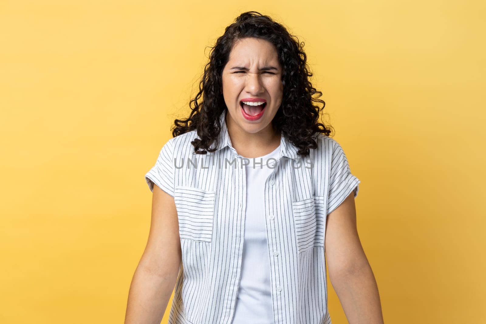 Portrait of angry aggressive young adult woman with dark wavy hair standing expressing aggression, screaming, frowning face. Indoor studio shot isolated on yellow background.