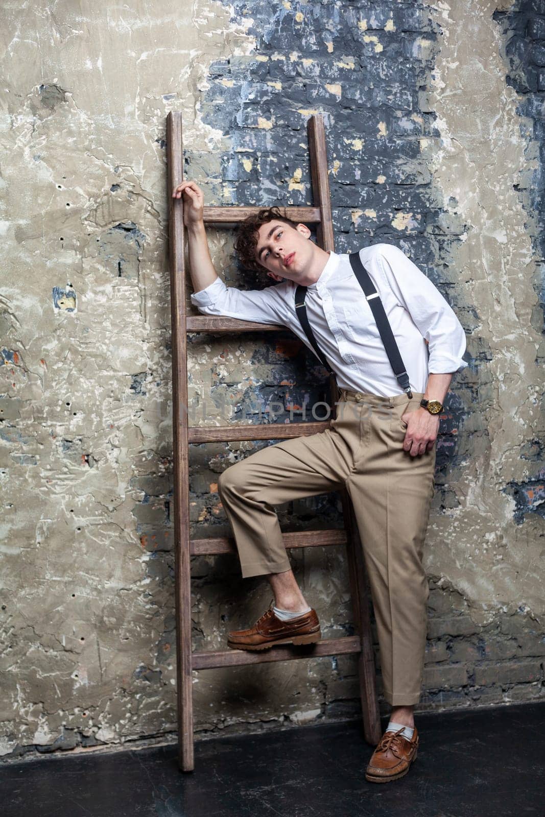 Portrait of trendy stylish young model man wearing white shirt and beige pants posing and lean on old wooden ladder on brick wall background. Indoor studio shot.
