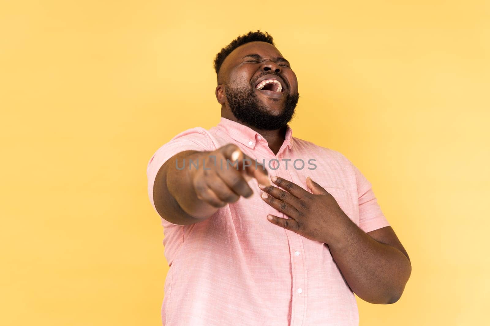 Portrait of funny positive bearded young adult man wearing pink shirt laughing out loud holding belly and pointing finger on you, mockery. Indoor studio shot isolated on yellow background.