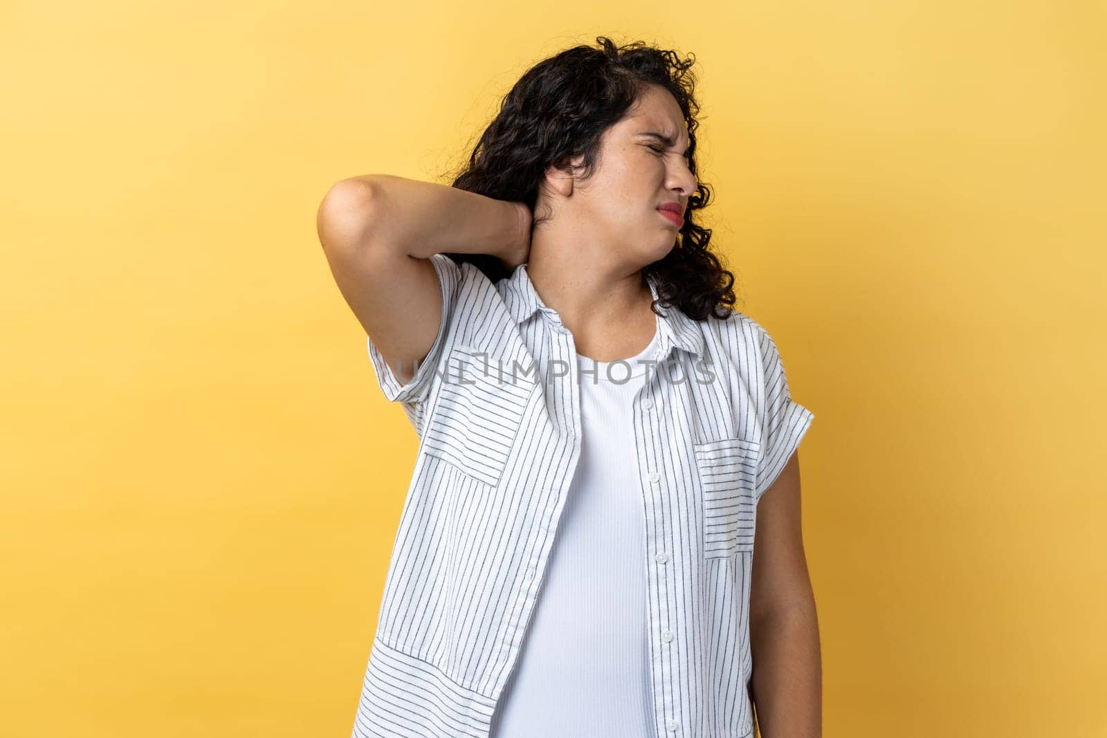 Portrait of woman with dark wavy hair touching neck, feeling acute pain, suffering spine problems, osteochondrosis, frowning face. Indoor studio shot isolated on yellow background.