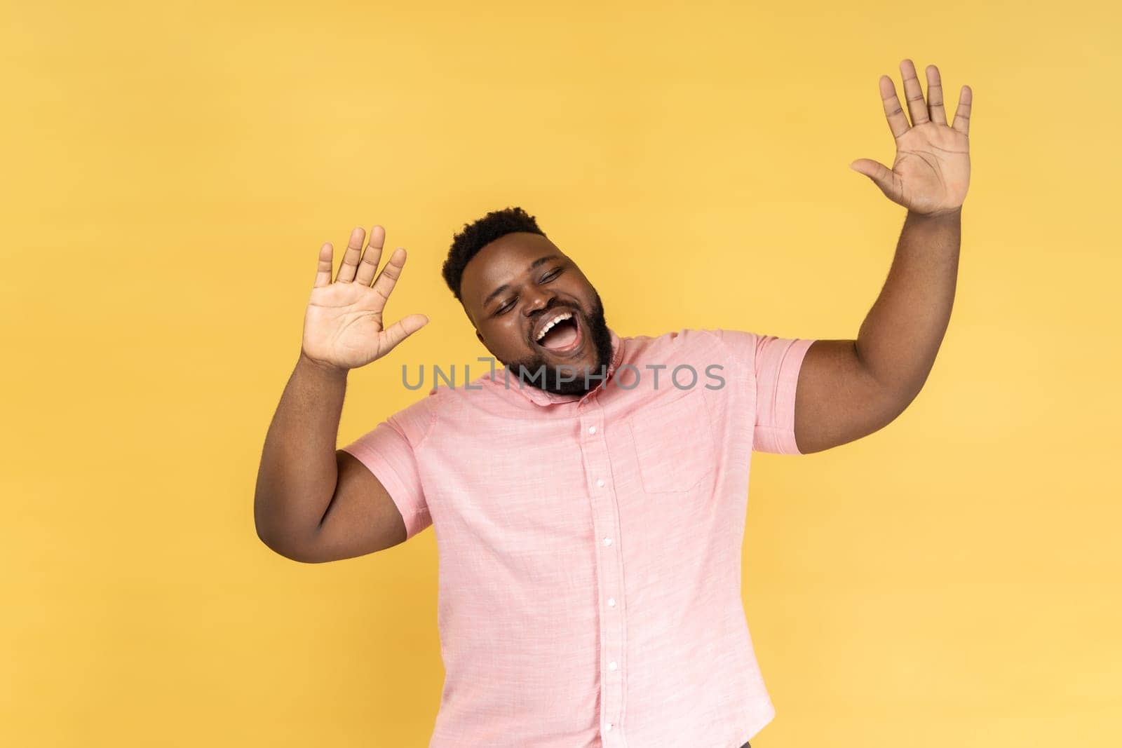 Portrait of excited positive man with beard wearing pink shirt dancing, raising hands up and having fun, being in good mood, celebrating. Indoor studio shot isolated on yellow background.