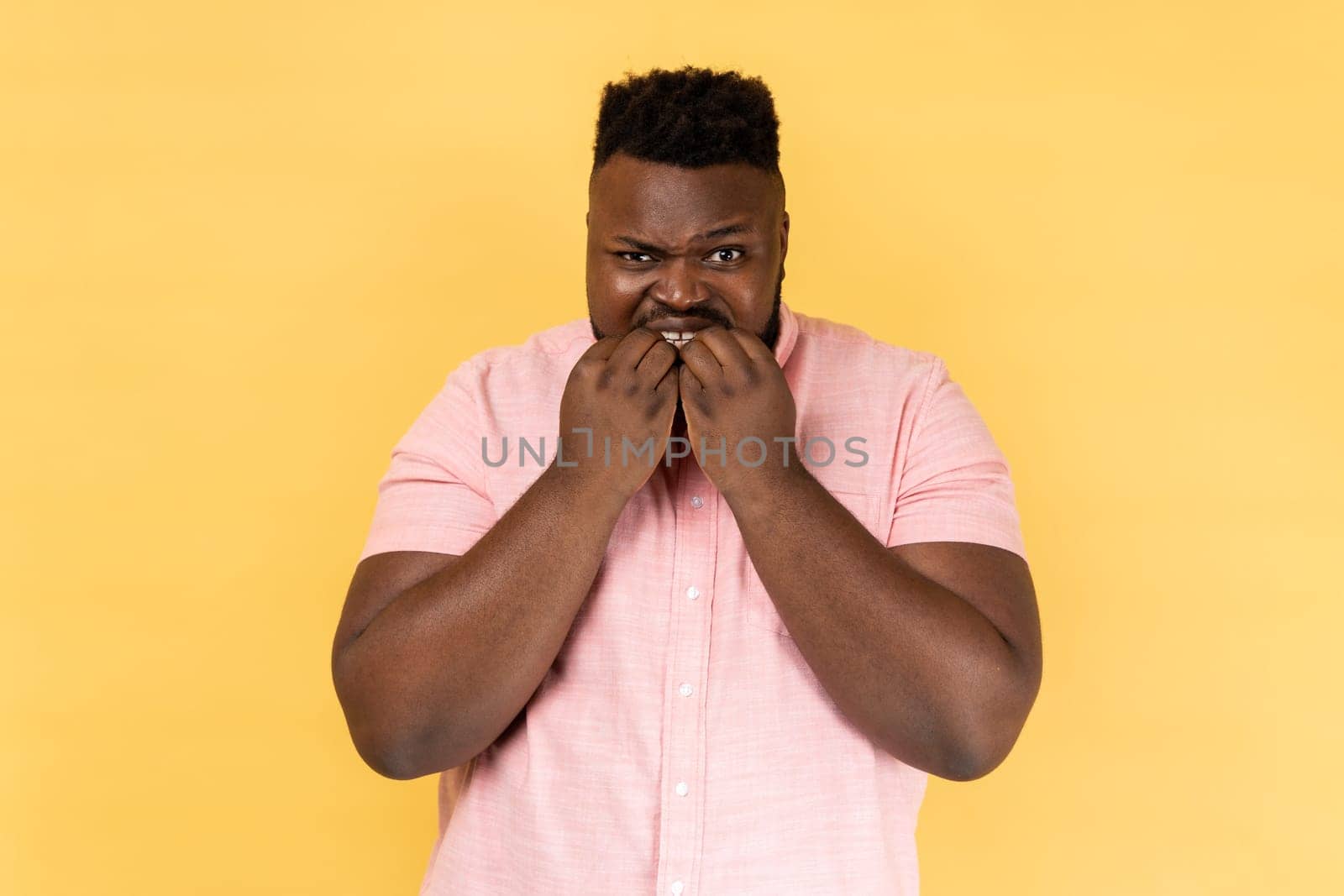 Portrait of panicking man in pink shirt biting nails, being nervous terrified, feeling frightened of challenge to start business, anxiety disorder. Indoor studio shot isolated on yellow background.