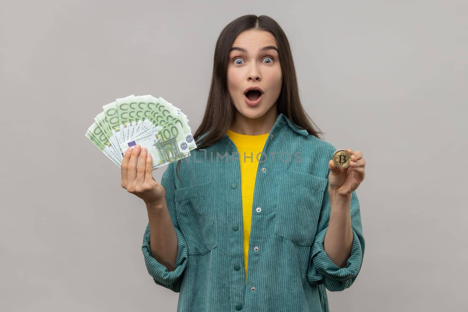 Portrait of excited shocked dark haired woman holding golden coin of crypto currency and big fan of euro banknotes, wearing casual style jacket. Indoor studio shot isolated on gray background.