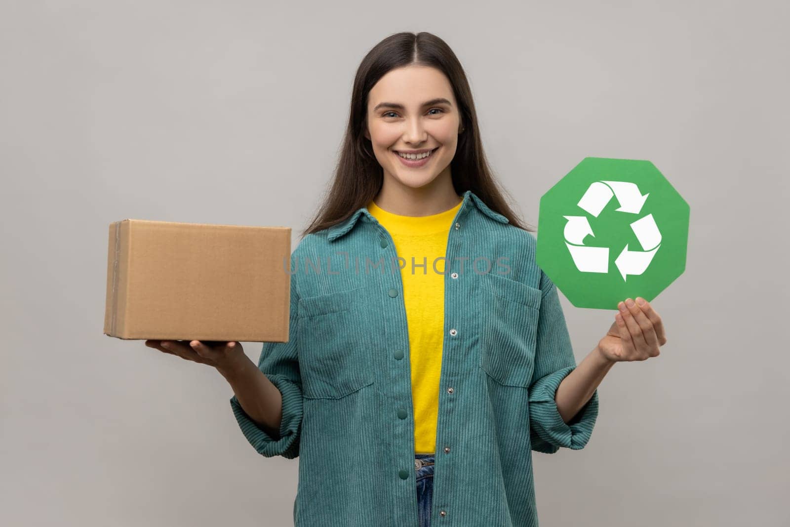 Happy positive young adult woman holding green recycling sign and cardboard package, saving environment, ecology concept, wearing casual style jacket. Indoor studio shot isolated on gray background.