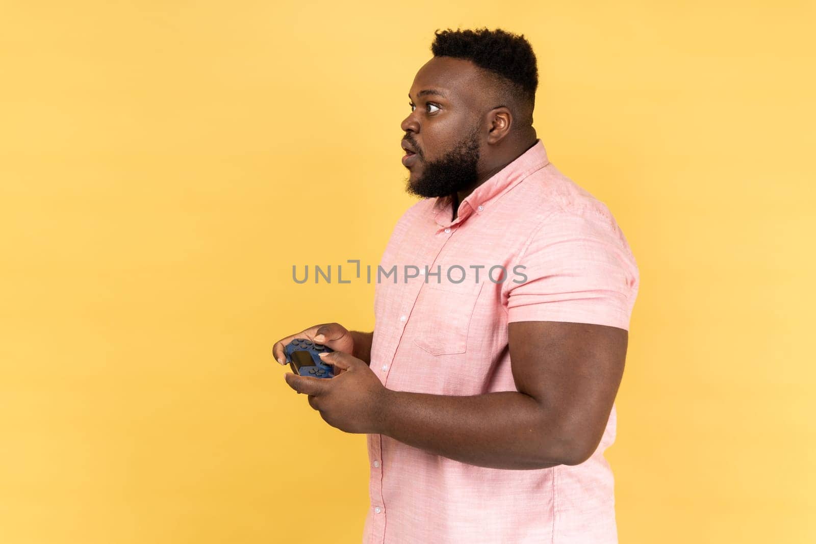 Side view of man wearing pink shirt holding in hands gamepad joystick, playing video games with big eyes and amazed facial expression. Indoor studio shot isolated on yellow background.