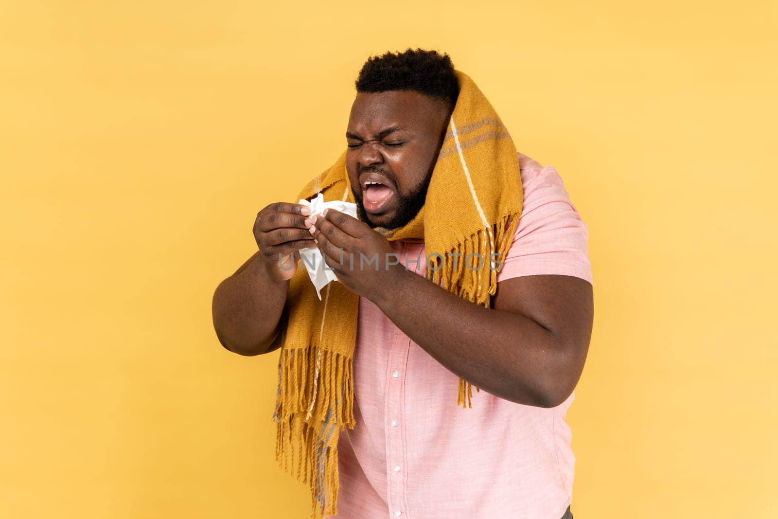 Portrait of unhealthy man in warm scarf coughing sneezing hard in napkin, feeling unwell with runny nose, severe seasonal allergy, influenza symptoms. Indoor studio shot isolated on yellow background.