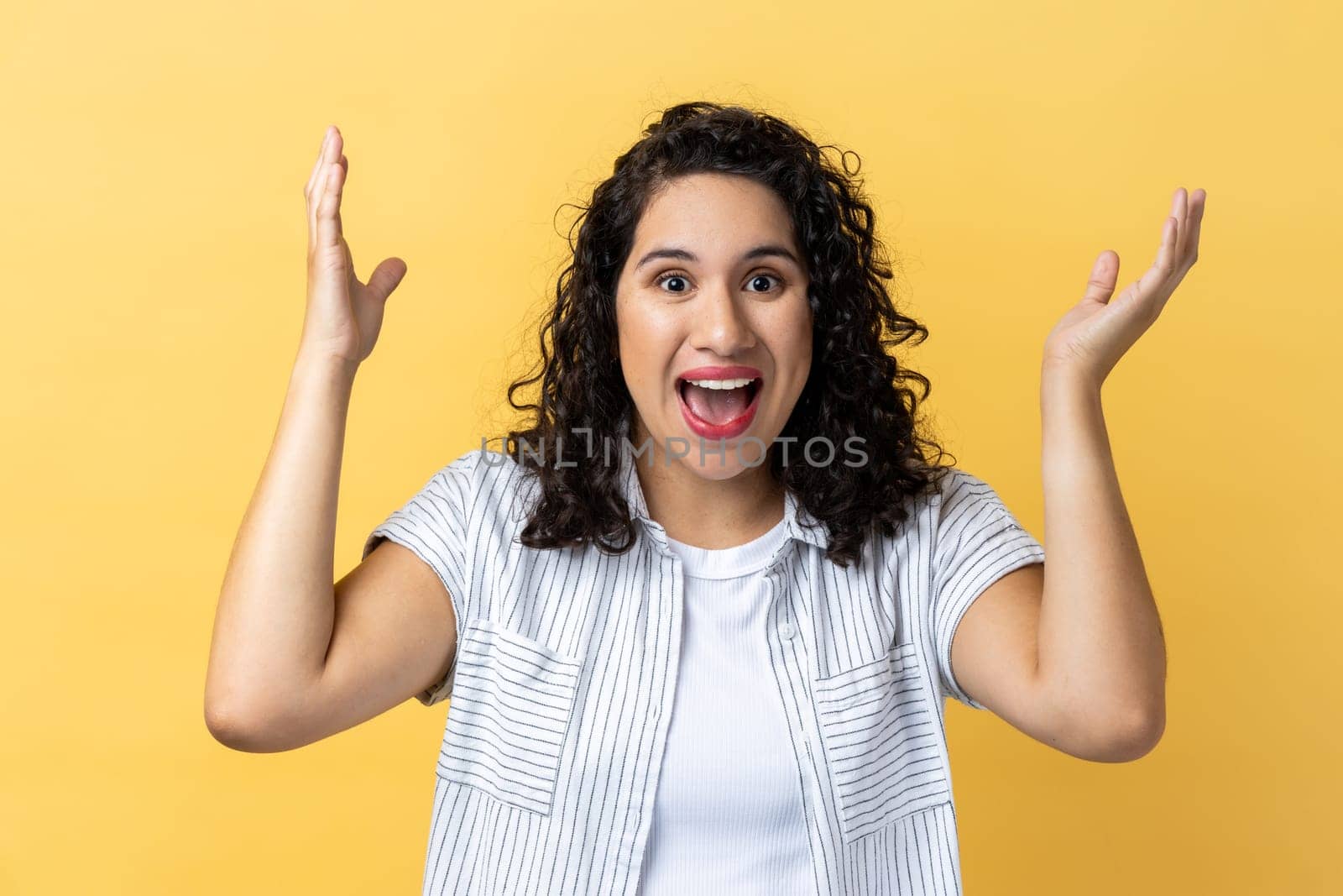 Portrait of amazed excited woman with dark wavy hair looking nat camera with excitement, keeping hands on cheeks, pleasant surprise. Indoor studio shot isolated on yellow background.