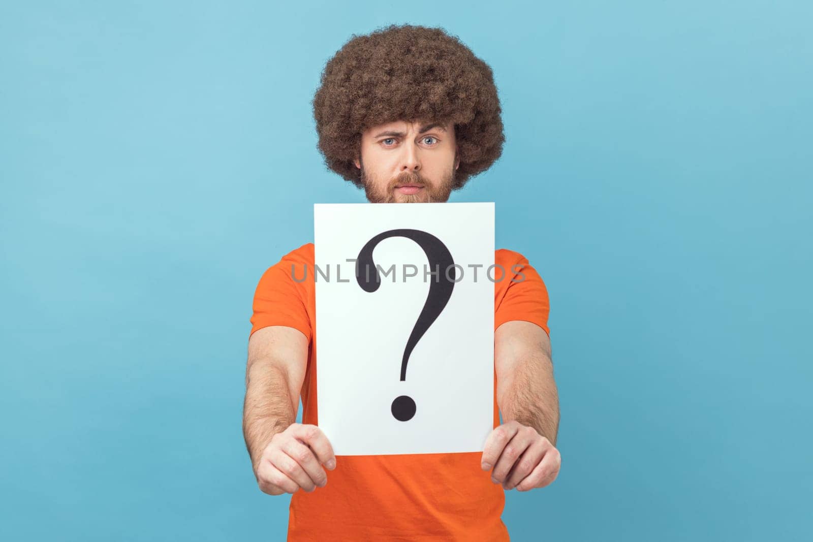 Portrait of man with Afro hairstyle wearing orange T-shirt holding question mark, finding smart solution, asking for advice, looking at camera. Indoor studio shot isolated on blue background.