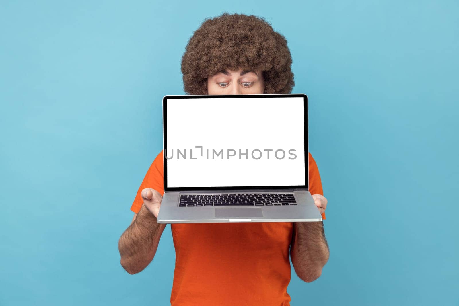 Portrait of unknown man with Afro hairstyle in orange T-shirt holding laptop in hands, hiding half of his face, copy space for promotion, looking down. Indoor studio shot isolated on blue background.