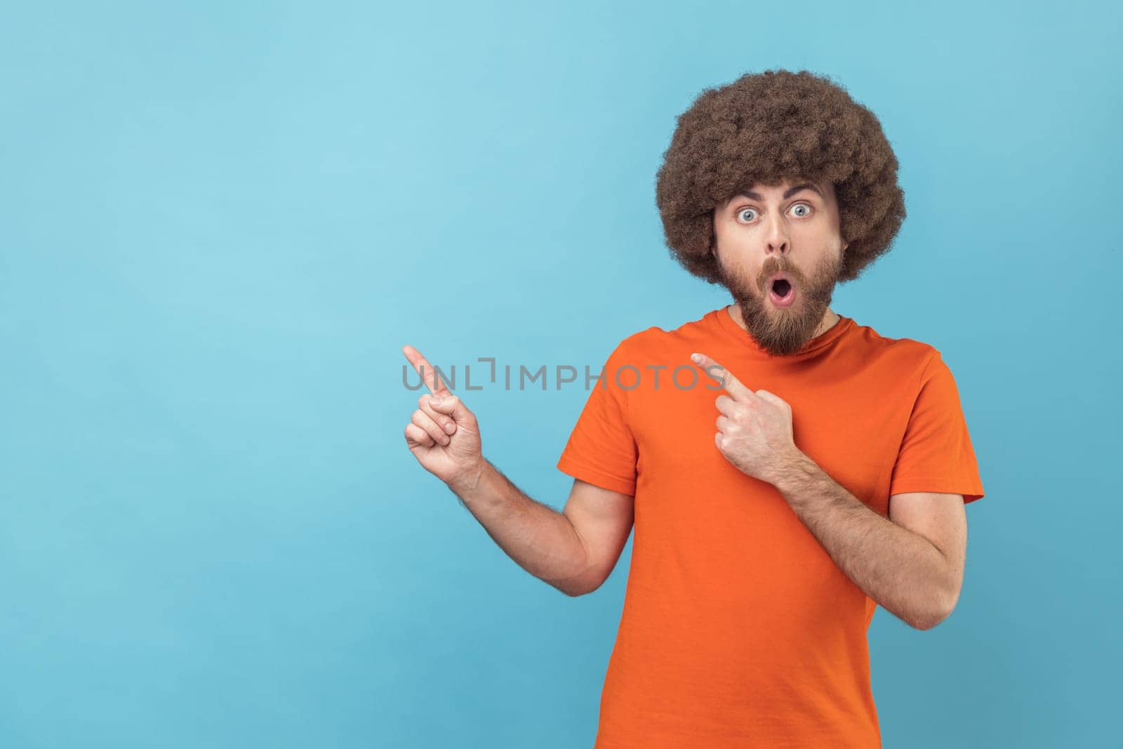Portrait of man with Afro hairstyle in orange T-shirt pointing aside, showing blank copy space for idea presentation, commercial text, looking at camera. Indoor studio shot isolated on blue background