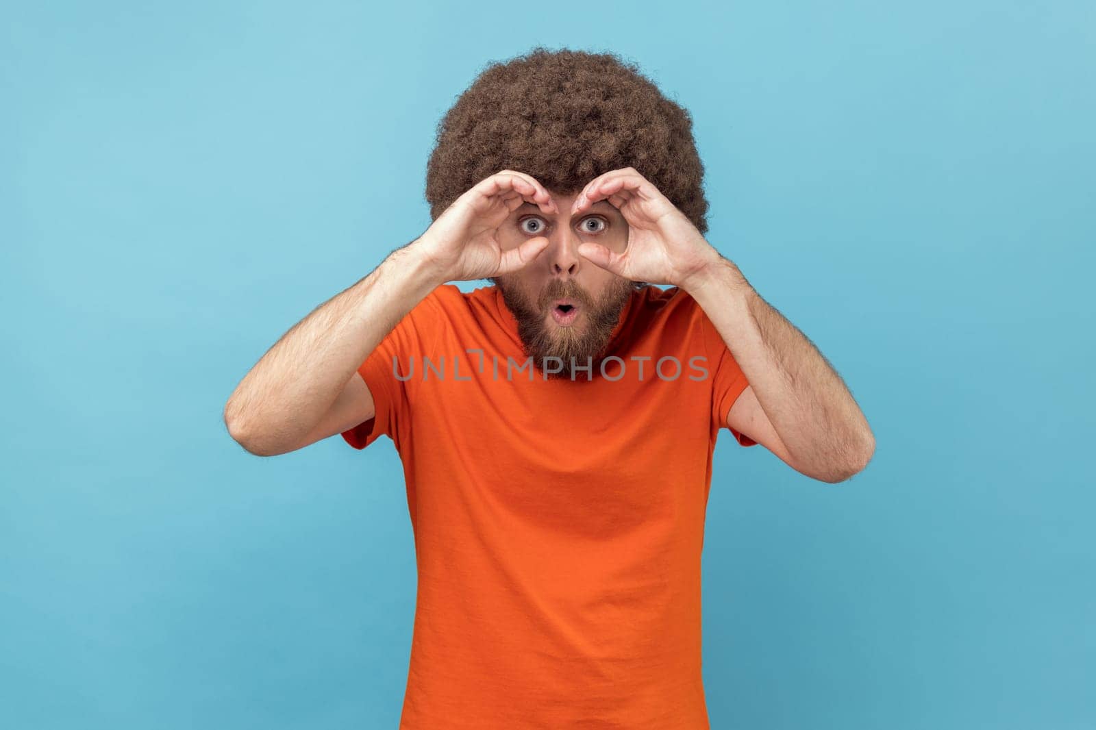 Portrait of man with Afro hairstyle wearing orange T-shirt holding fists near eyes imagining binoculars and looking through holes, spying, having fun. Indoor studio shot isolated on blue background.