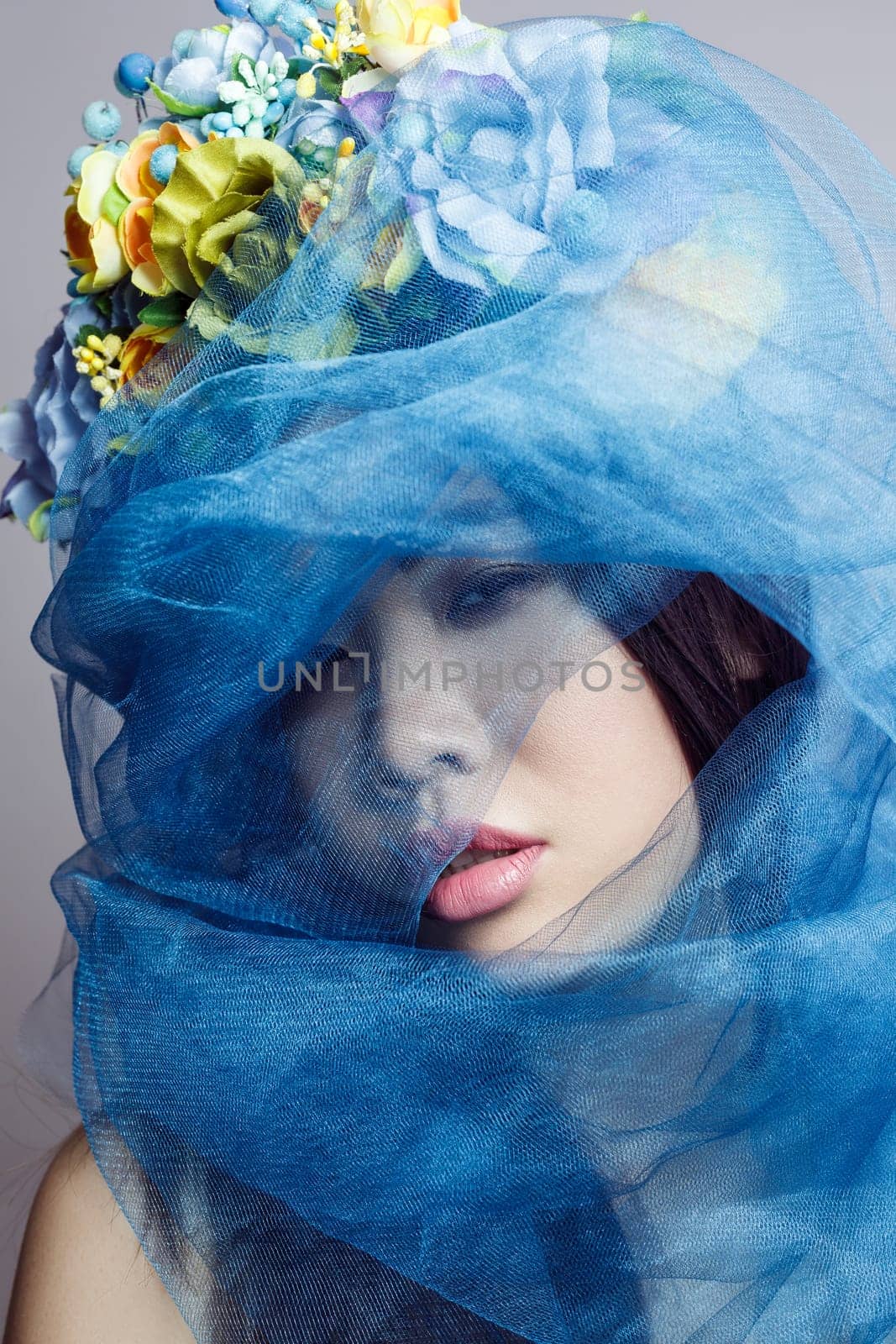 Closeup portrait of mysterious attractive sensual beautiful woman with floral hat, female with makeup, hiding her face with blue veil. Indoor studio shot isolated on gray background,