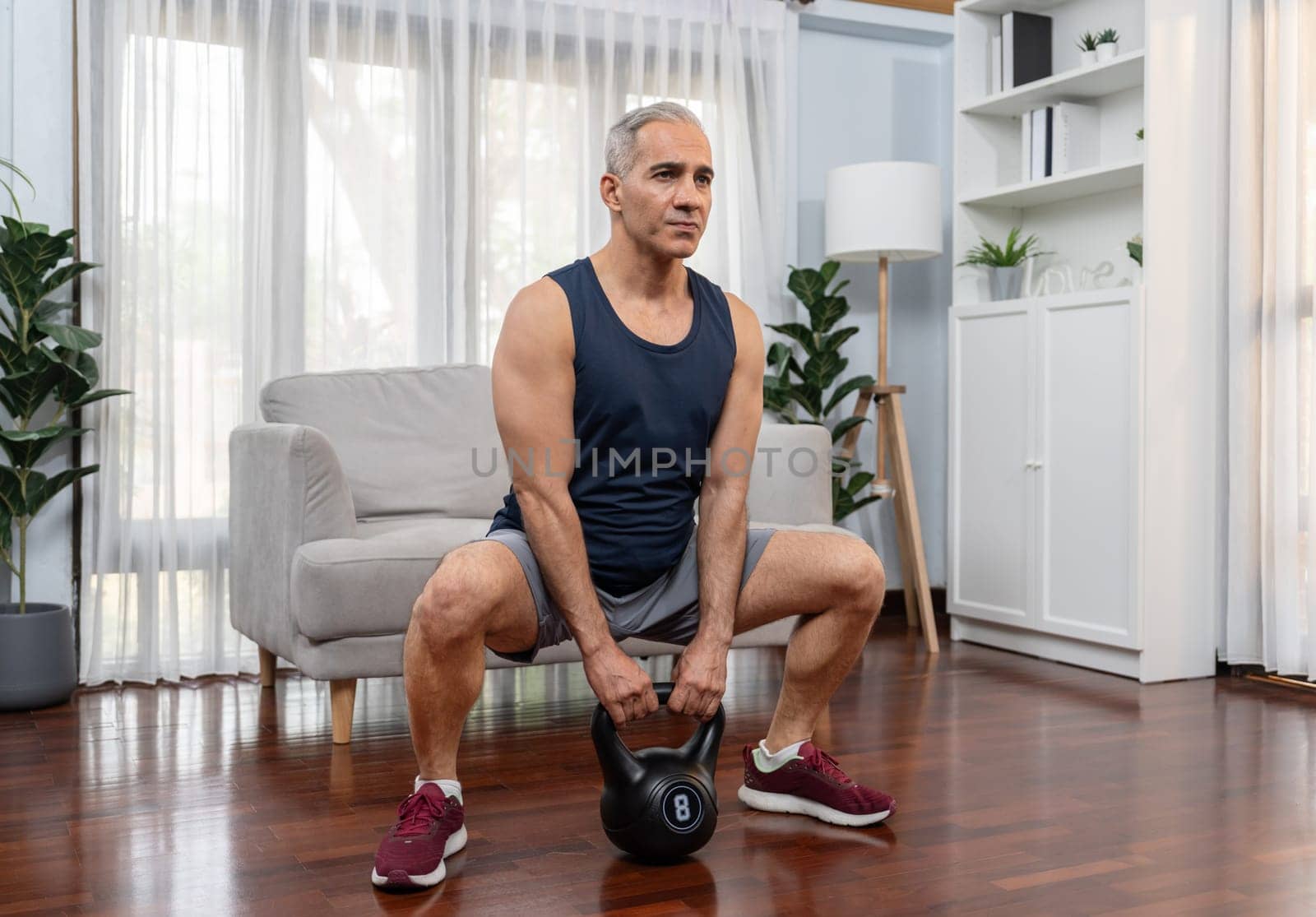 Athletic and sporty senior man engaging in leg day training session with squat and bodyweight kettle ball at home exercise as concept of healthy fit body lifestyle after retirement. Clout