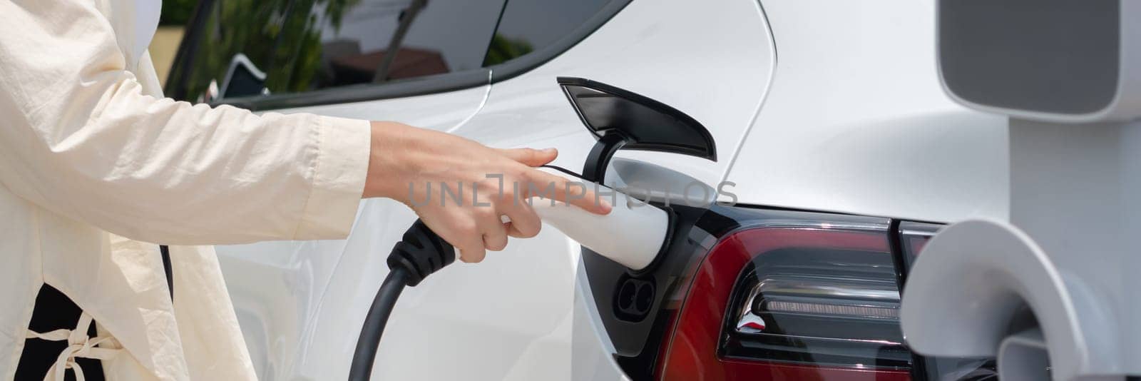 Young woman put EV charger to recharge electric car's battery from charging station. Alternative energy and rechargeable EV car for sustainable environmental friendly travel concept.Panorama Expedient