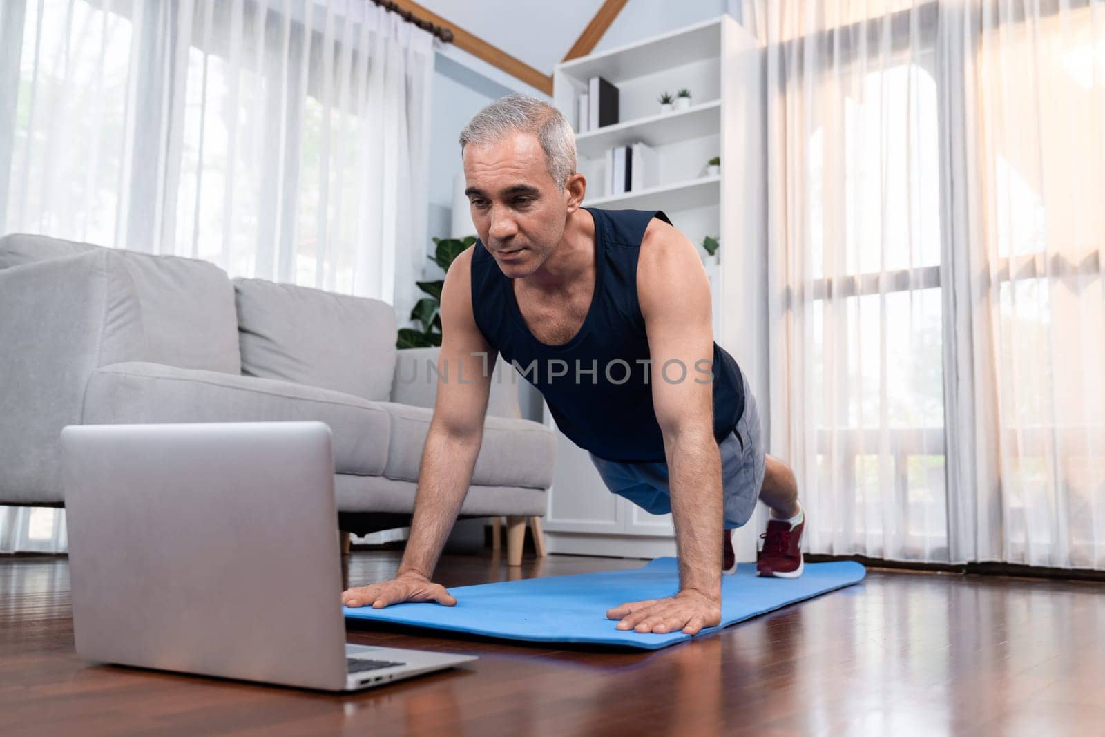 Athletic and sporty senior man doing pushup on exercising mat while watching instruction from online video at home exercise as concept of healthy fit body lifestyle after retirement. Clout