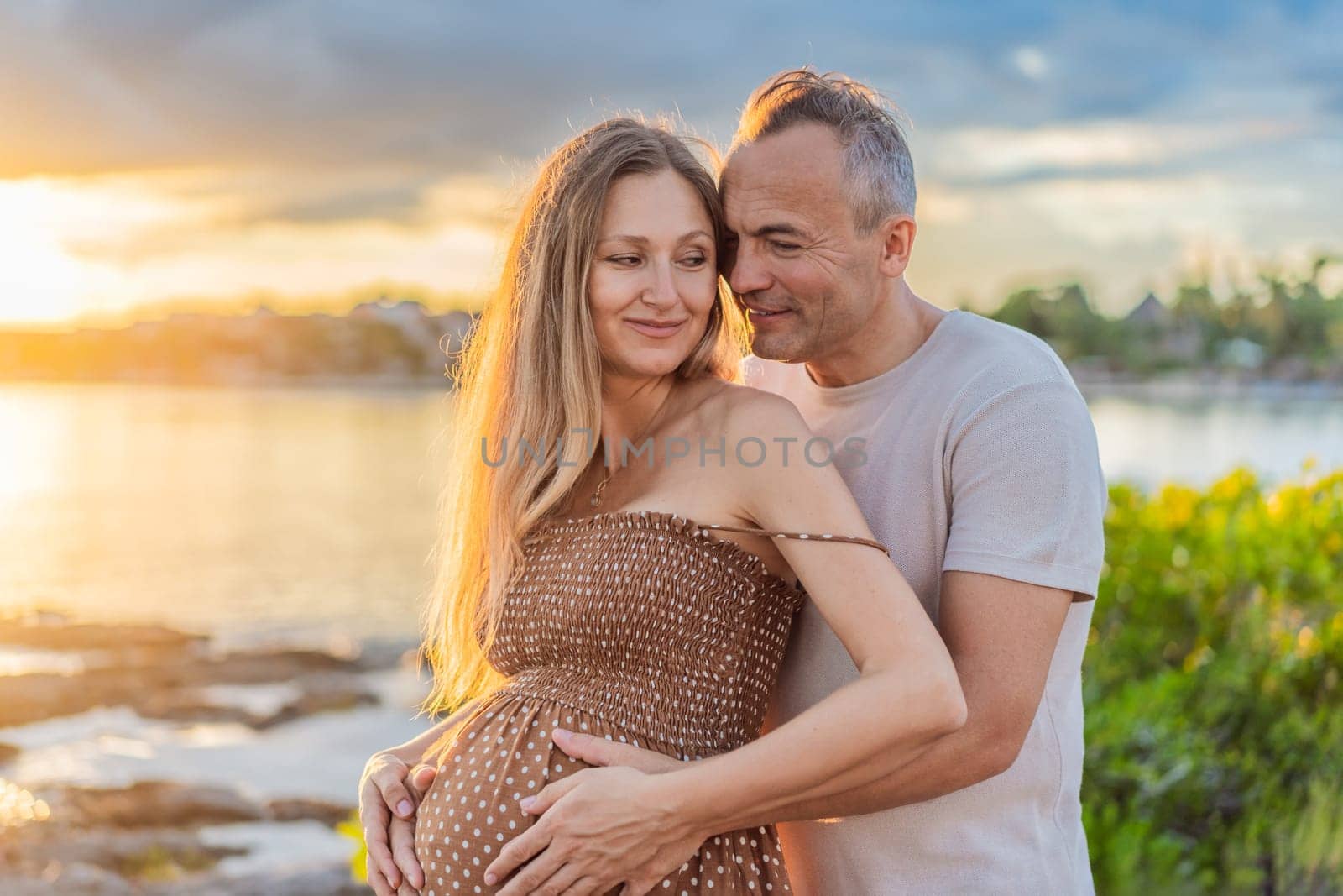 A happy, mature couple over 40, enjoying a leisurely walk on the waterfront On the Sunset, their joy evident as they embrace the journey of pregnancy later in life by galitskaya