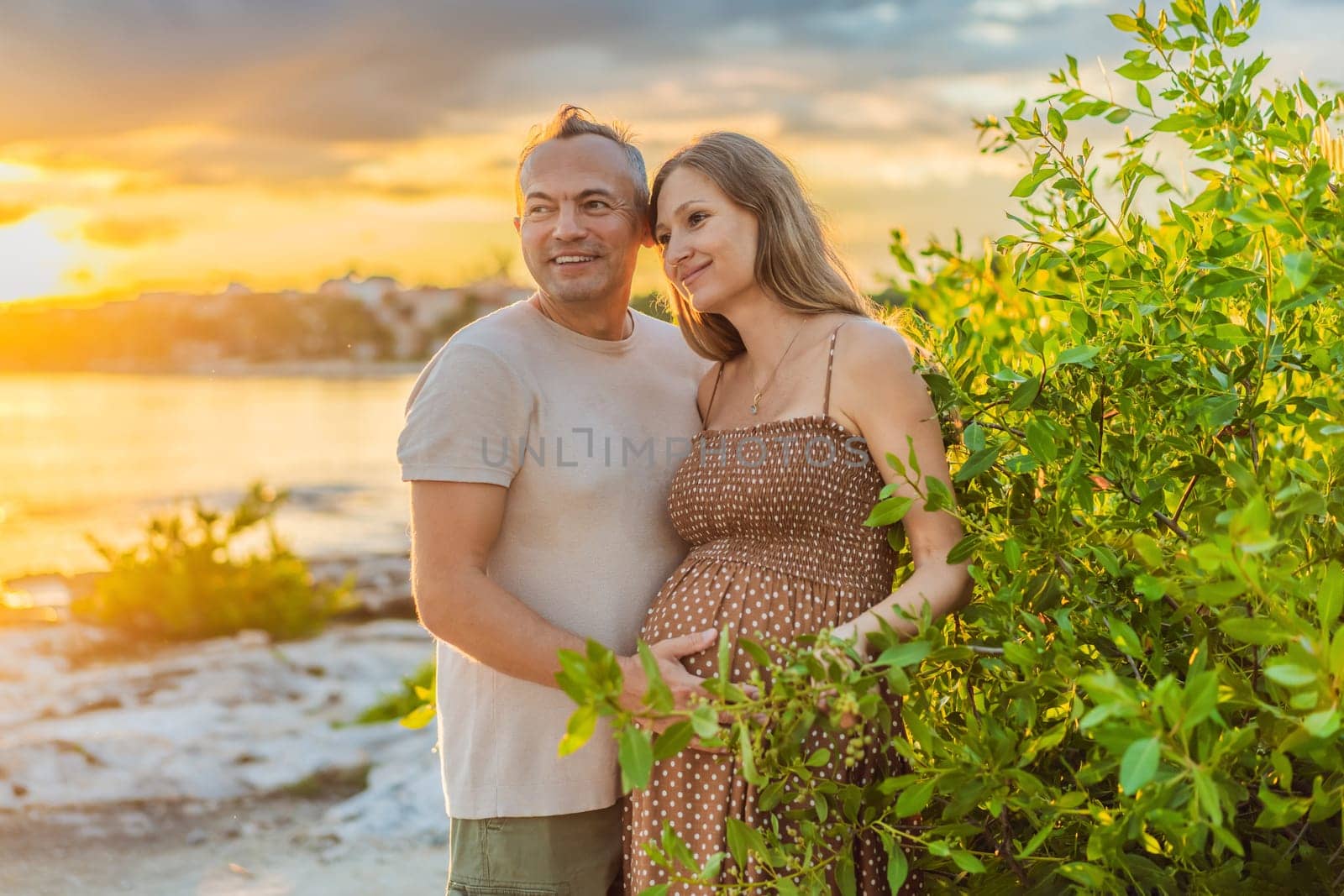 A happy, mature couple over 40, enjoying a leisurely walk on the waterfront On the Sunset, their joy evident as they embrace the journey of pregnancy later in life by galitskaya